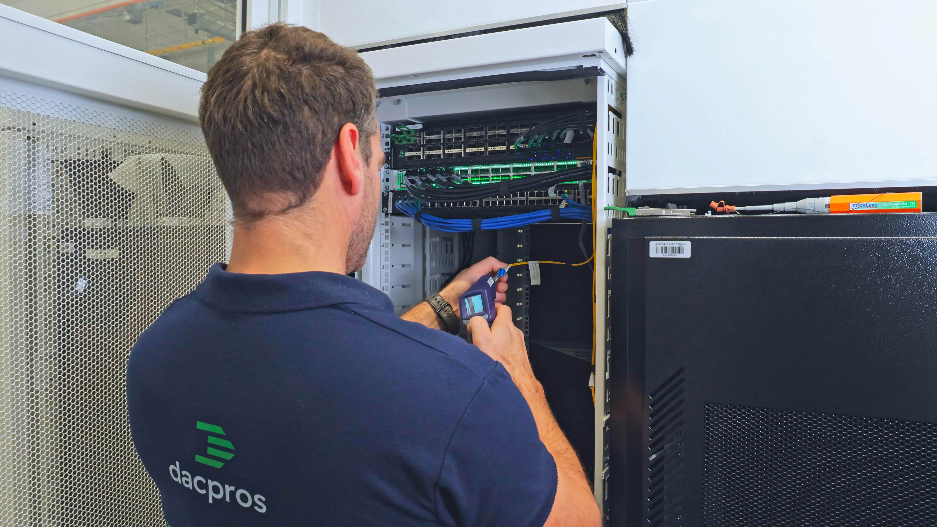 DACPROS engineer using an OTDR testing device at an open comms cabinet — verification and planning in progress.