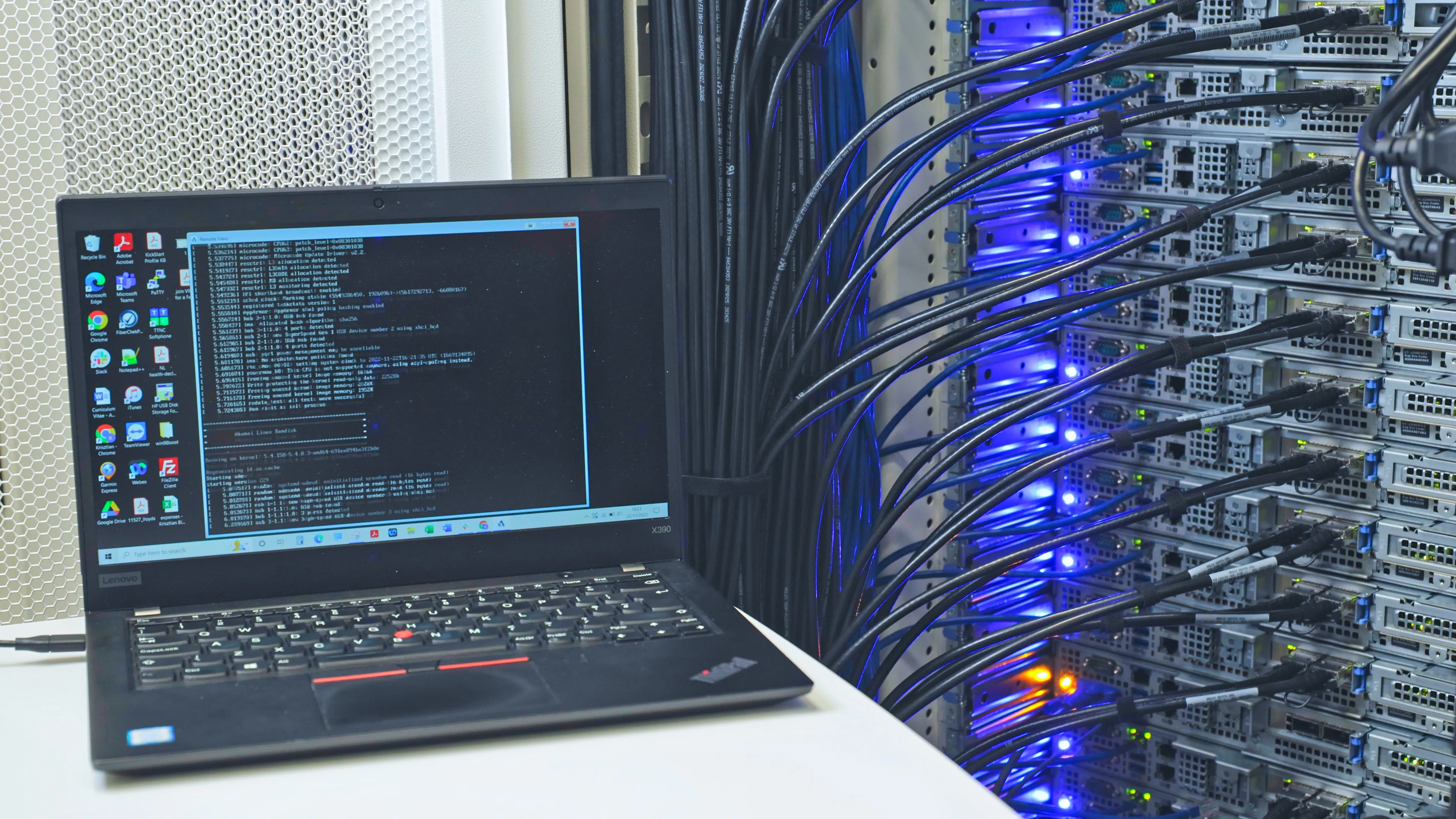 Laptop running terminal diagnostics next to a densely cabled rack of servers with blue LED status lights — post-deployment verification.